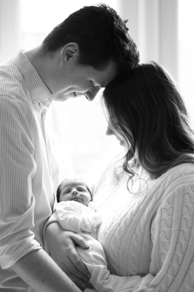 Parents smile down at their newborn as the light from the window behind shines in.
