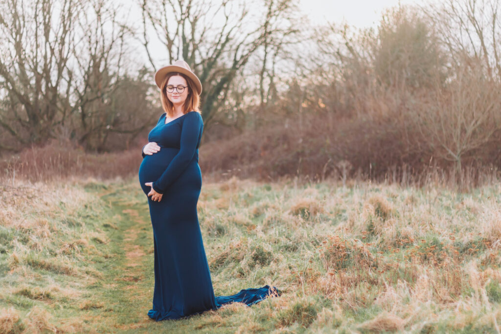 Mum-to-be poses in a blue floor length dress and brown hat standing in a Somerset park