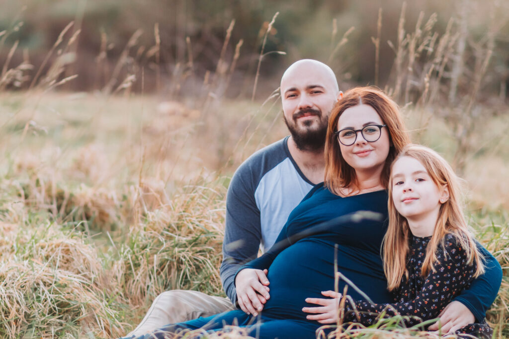 Family of three smile at he camera as they sit in the long grass cuddled around Mum-to-be.