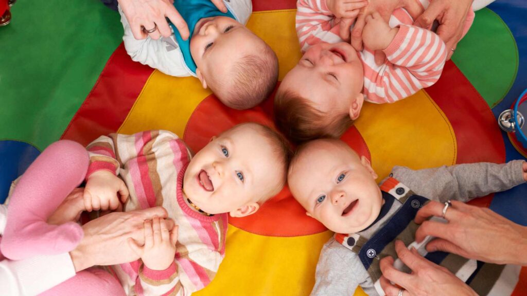 Four babies laying on colourful blanket being tickled by their adult