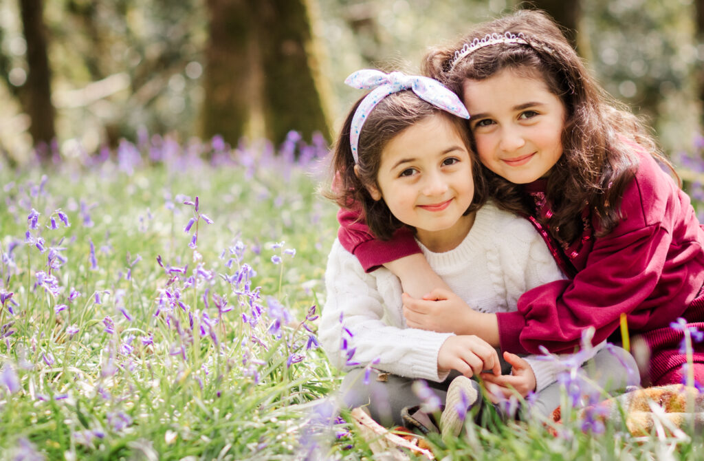 Two young sisters sit amongst the bluebells smiling and hugging for the camera