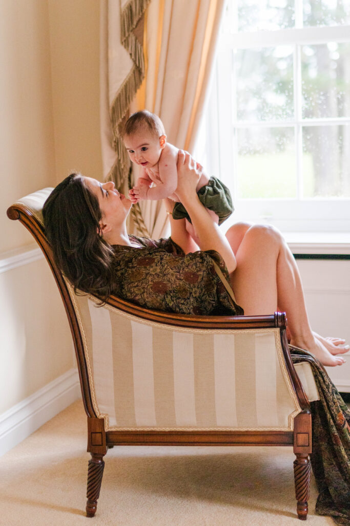 Mum holds her baby up and laughs as she sits in a chair by the window