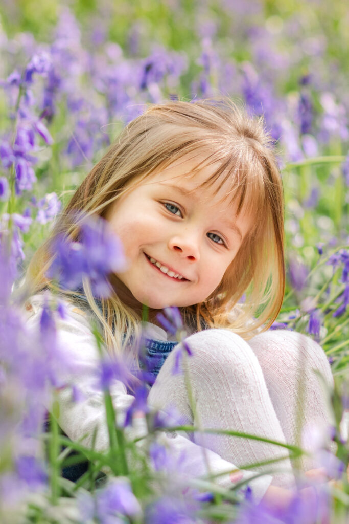 Little girl smiles as she sits surrounded by a sea of bluebells