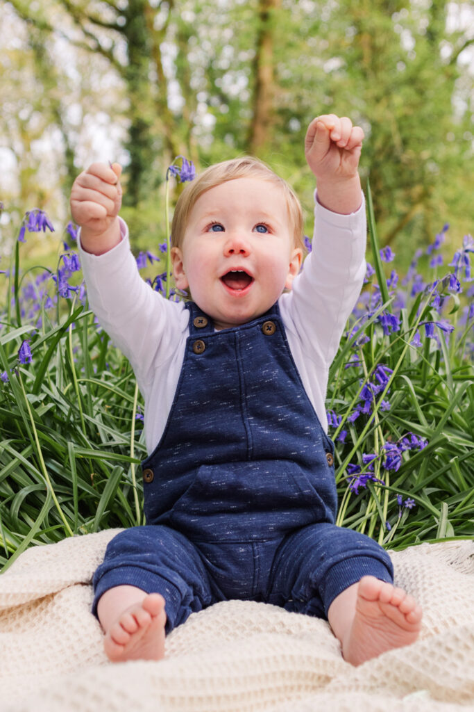 Little boy sits raises his arms up in glee as he sits on a blanket in front of some bluebells