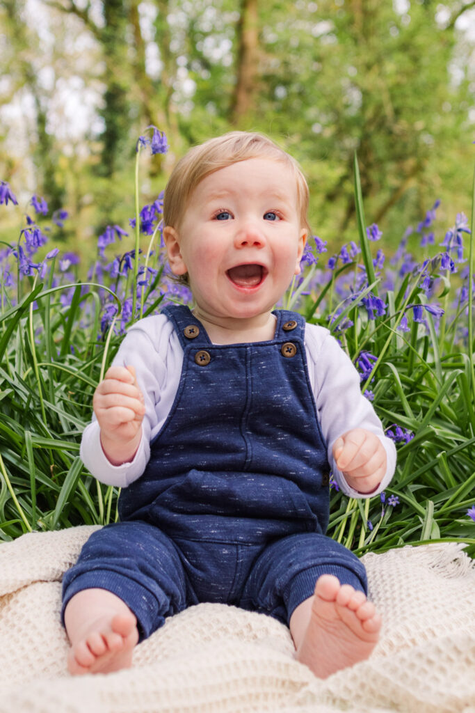Little boy smiling as he sits on a blanket in front of bluebells 