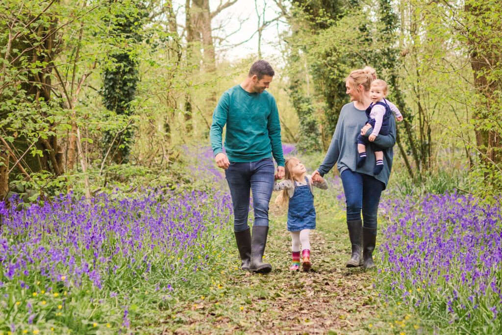 Family of four walk together along a woodland path sided by bluebells
