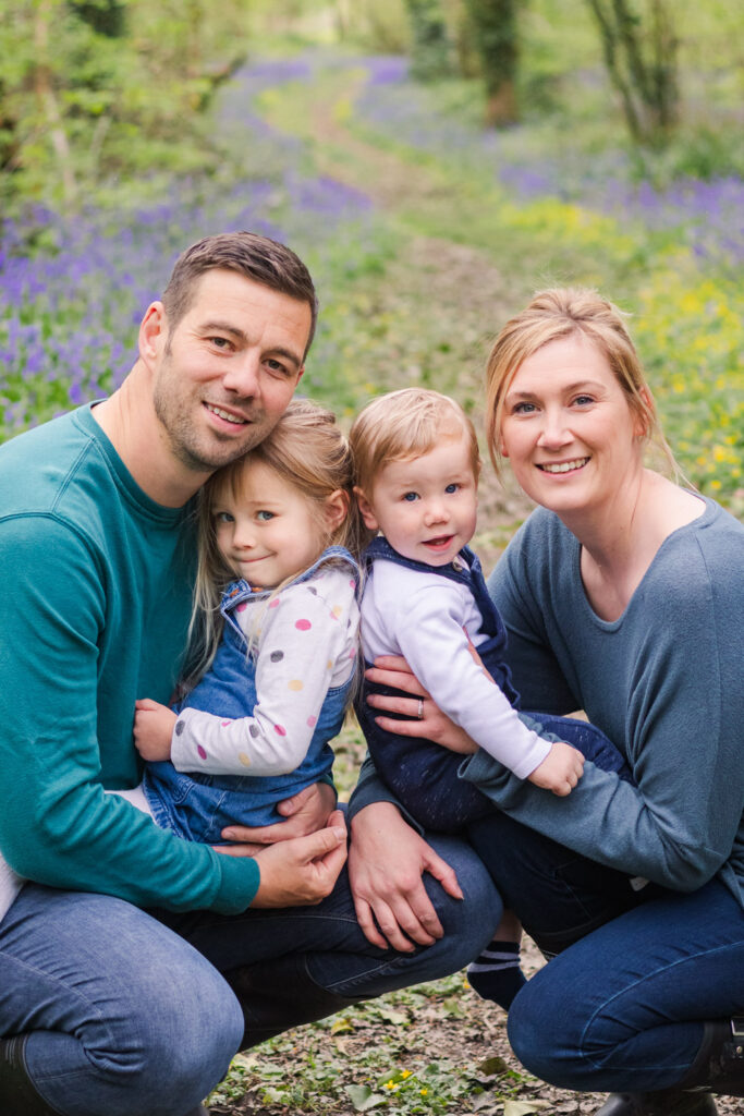Family pose together for a photograph in front of a bluebell lined path
