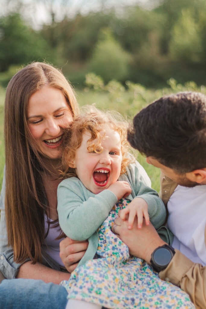 Little girl is tickled by her parents as she looks towards the photographer