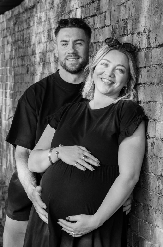 Couple lean against a wall smiling towards the camera as they cradle their baby bump