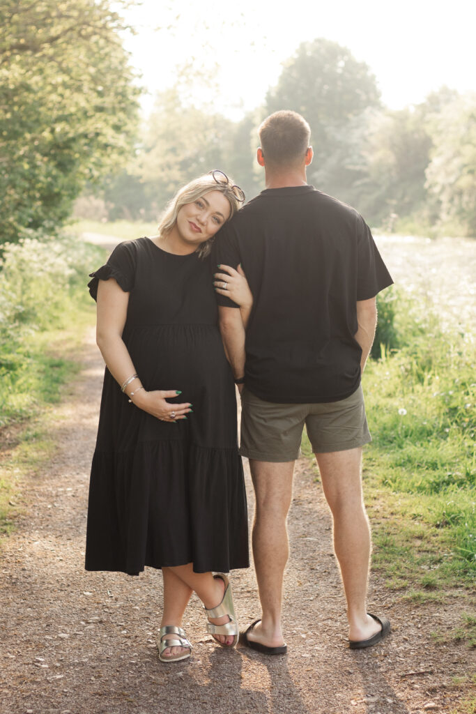 Pregnant lady looks at camera and leans on her husbands shoulder as he looks away at the view