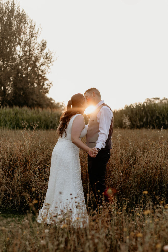 Bride and groom share a kiss in flower meadow as the sun sets behind them