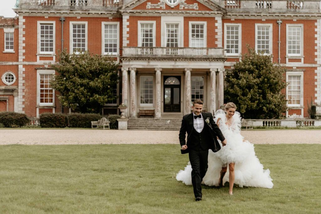 Bride and groom holding hands, run across the grass in front of their Country House Wedding Venue