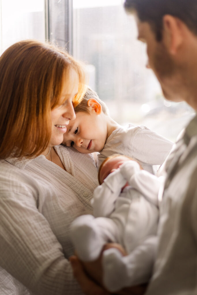 Family with newborn stand in front of the window cuddling together