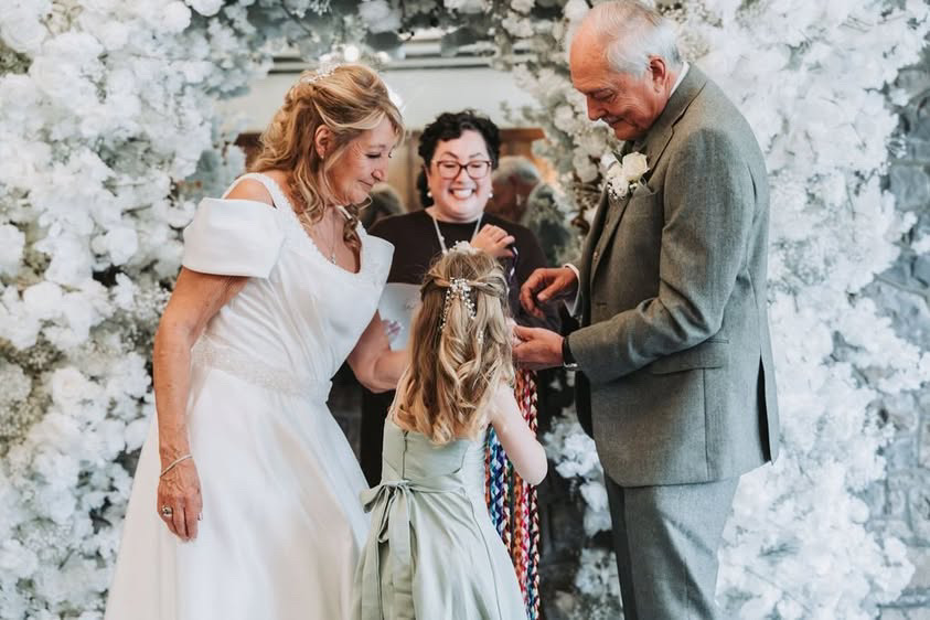Ring bearer presents rings to wedding couple as smiling wedding celebrant looks on